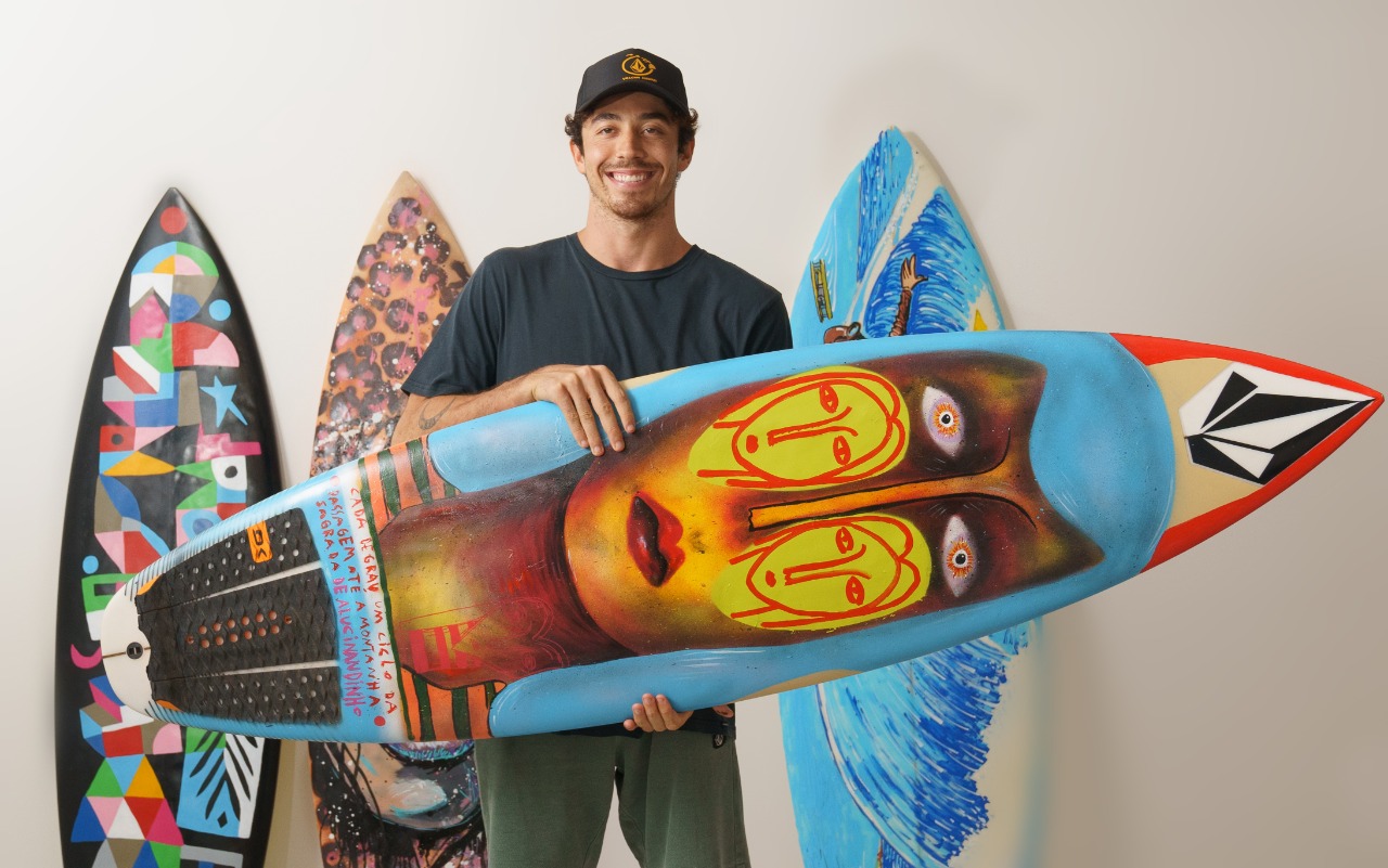 O surfista Yago Dora segurando sua prancha pintada pelo artista Alucinandinho -
